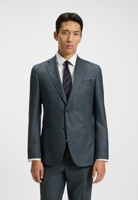 Op maat gemaakt donkerblauw pak in een gestructureerde stof, met een enkelvoudige sluiting, geknipte revers, gecombineerd met een wit overhemd en een gestreepte das, inclusief een pochet.
