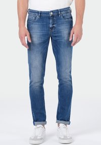 Jean en denim bleu à coupe slim, présentant des motifs délavés, cinq poches et une braguette éclair. À assortir avec des baskets blanches.