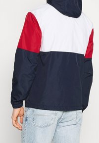 Färgglad vindjacka med paneler i marinblått, rött och vitt. Tillverkad av lätt, glänsande material med elastiska ärmslut och huva.