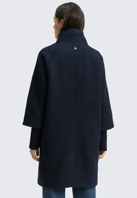 Dunkelblauer Fleece-Mantel mit hohem Kragen, kurzen Ärmeln und strukturierten Oberflächen. Verfügt über einen einzelnen Knopfdetail auf der Rückseite.