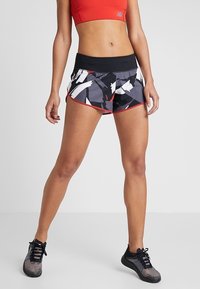 Kvinna står och bär en röd sport-bh, svart och vit mönstrad löpshorts med röd kant, samt svarta träningsskor på vit bakgrund.