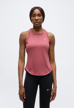 Žena oblečená v ružovom tielku Nike bez rukávov a čiernych legínach Nike, stojaca pred jednofarebným svetlým pozadím.