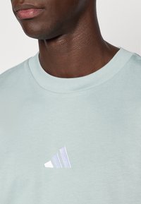 Gaiši zaļa kokvilnas T-krekls ar apaļu kakla izgriezumu un izšūtu baltu un violetu Adidas logotipu kreisajā krūtīs.