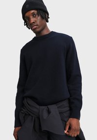 Marineblauer Strickpullover mit Rundhalsausschnitt und langen Ärmeln. Gerippte Bündchen und Saum. Getragen über hoch Taillierten dunklen Hosen mit gebundenem Bund.