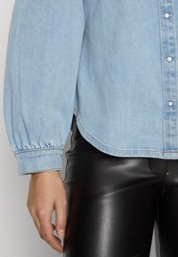 Lys blå denimskjorte med puffede lange ermer og knappestolpe. Skjorten har subtile sømdetaljer og en buet kant. Kombinert med svarte skinnbukser.