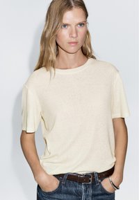 Světlý žlutý tričko s krátkým rukávem a kulatým výstřihem, s mírnou texturou, v kombinaci s tmavými džínami a hnědým páskem. Jednoduchý design.