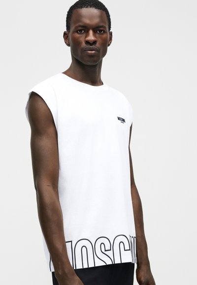 Άνδρας που φοράει λευκό αμάνικο μπλουζάκι με μαύρο κείμενο "MOSCHINO" στο στήθος και μεγάλα περιγραμμένα γράμματα κατά μήκος του τελειώματος, στέκεται μπροστά σε απλό φόντο.