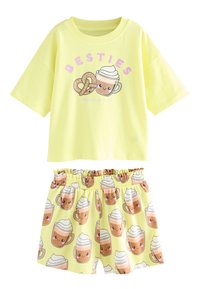 Kollane T-särk üle suuruse lõikega ja mängulise kohvitassi ja kringli graafikaga. Sobivad lühikesed püksid on kaetud korduva kohvitassi printidega.