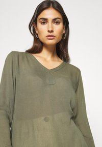 Long-sleeve olive green blouse with a V-neckline, featuring a textured fabric and a small front placket detail.