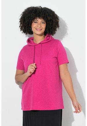 Smilende kvinde med krøllet hår iført en lyserød kortærmet hættetrøje og sort nederdel, stående mod en hvid baggrund.
