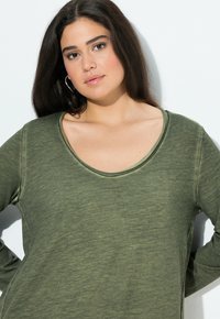 Jonge vrouw met lang, donker haar in een groene lange mouwen top, staande tegen een witte achtergrond, zacht kijkend naar de camera.