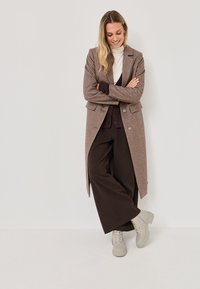 Abrigo largo de mezcla de lana marrón con corte entallado, botones negros, superpuesto a un suéter de cuello alto crema y pantalones oscuros, combinado con zapatos gris claro.