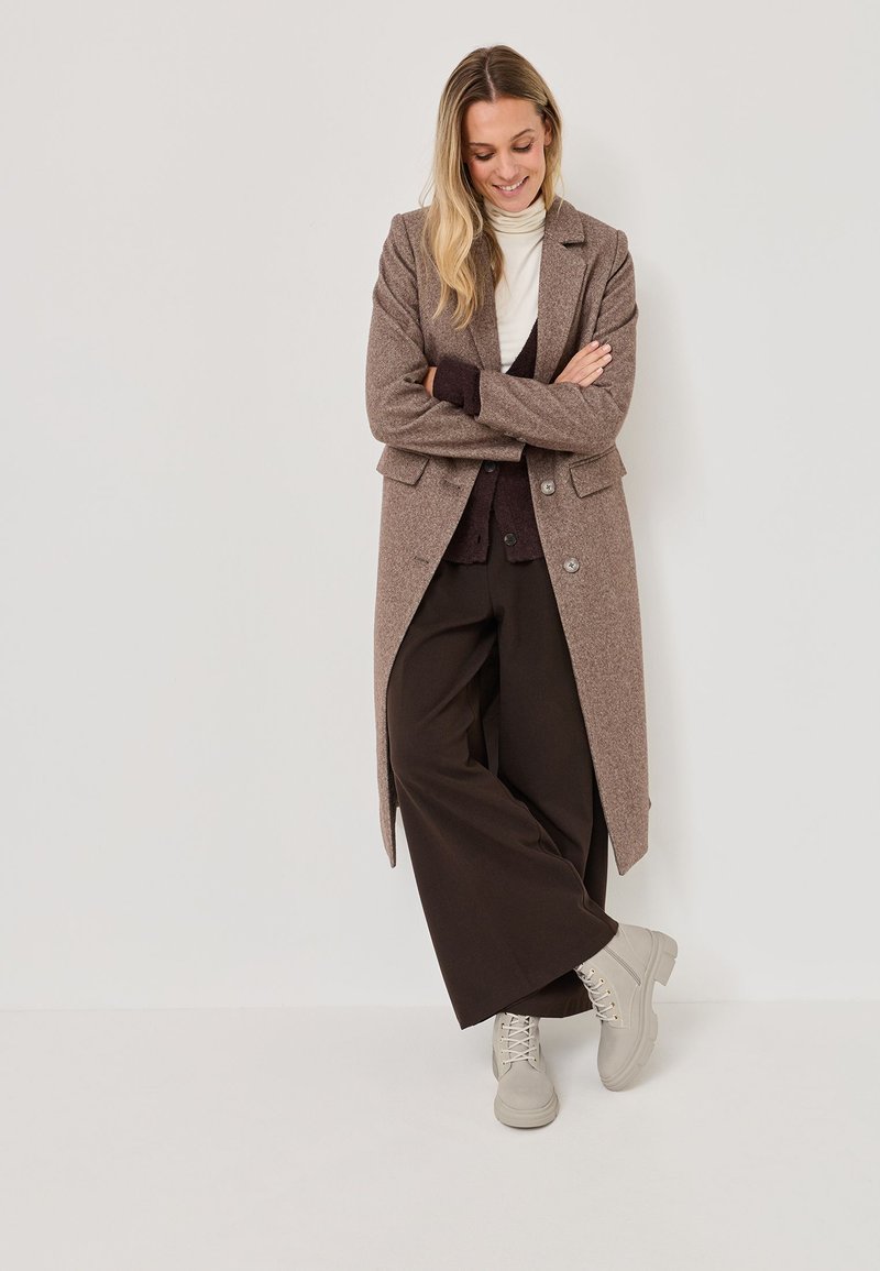 Abrigo largo de mezcla de lana marrón con corte entallado, botones negros, superpuesto a un suéter de cuello alto crema y pantalones oscuros, combinado con zapatos gris claro.