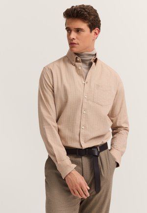 Ein Mann trägt ein beiges Hemd mit Knöpfen über einem hellbraunen Rollkragenpullover, braune Hose und einen schwarzen Gürtel. Er schaut zur Seite, während eine Hand in der Tasche ist.