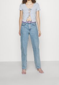 Žena v světle modrém zkráceném topu s předními vázáními, v volných modrých rifle s vysokým pasem, s fialovým páskem a levandulovými sandály na podpatku se stahovacími pásky.