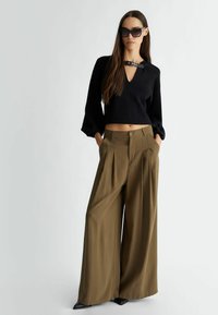 Maglione nero cropped con scollo a V e un dettaglio in pelle al colletto, abbinato a pantaloni a vita alta plissettati verde oliva e scarpe nere a punta.