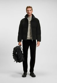 Sort puffjakke med høj krave, grå striktrøje, sorte fløjl bukser og robuste sorte sneakers. Bærer en sort rygsæk.