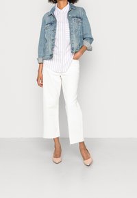 Veste en denim bleu clair, chemise blanche à rayures, jean blanc à jambes larges avec ourlet effiloché, et talons hauts couleur nude. Tenue décontractée en superposition.