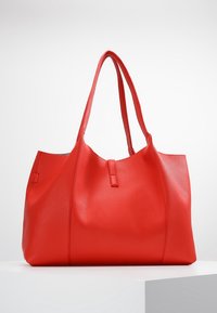 Punainen nahkainen kassi, jossa on teksturoitu pinta, varustettu kahtena pitkänä kahvana ja avoimella yläsuunnalla, esillä neutraalilla taustalla.