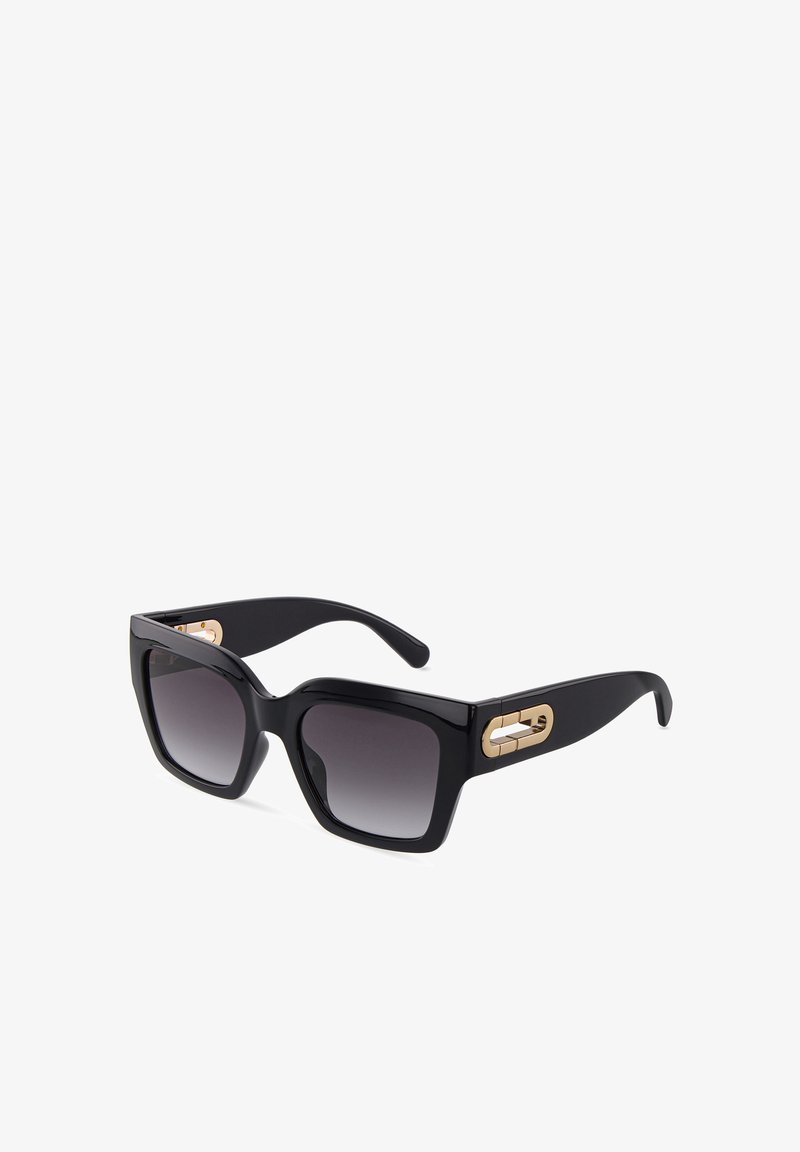 Lunettes de soleil carrées noires oversize avec verres foncés dégradés et accents décoratifs dorés sur les branches sur fond blanc.