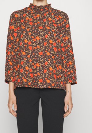 Kvinde iført en brun bluse med lange ærmer med orange blomsterprint og flæsekrave sammen med sorte bukser.