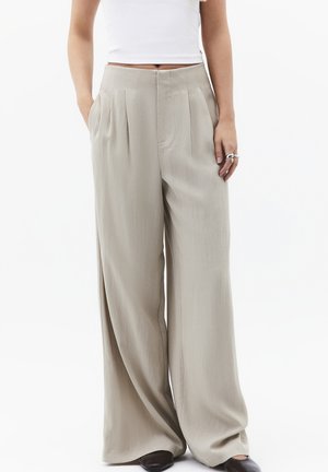 Donna che indossa pantaloni beige a vita alta e gamba larga con pieghe frontali, abbinati a un crop top bianco e scarpe nere.