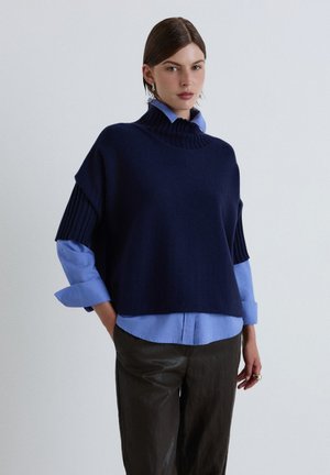 Donna che indossa un maglione a maniche corte blu navy sopra una camicia azzurra con maniche arrotolate e pantaloni neri, in piedi davanti a uno sfondo uniforme.