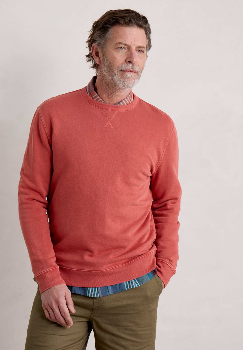 Felpa rossa in cotone con scollatura a girocollo, caratterizzata da una texture semplice. Indossata sopra una camicia a righe blu e bianche, abbinata a pantaloni beige.