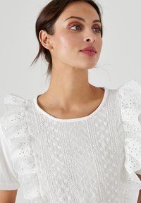 Blusa bianca con maniche in pizzo e ricami intricati. Il tessuto ha una consistenza morbida con dettagli traforati decorativi e un collo rotondo.