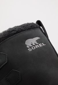 Botte d'hiver noire avec une doublure en polaire douce et un logo argenté Sorel représentant un ours sur le dessus.
