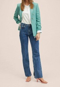 Femme portant un blazer bleu sarcelle avec des boutons dorés, un chemisier blanc, un jean taille haute bleu avec des fentes à l'avant, et des sandales à talons roses, debout avec une main dans la poche.