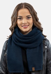 Eșarfă tricotată de culoare bleu marin, cu o suprafață texturată, înfășurată în jurul gâtului, având un mic etichetă din piele cu brand pe lateral.