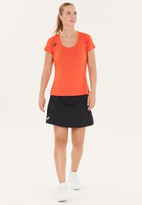 Orange, kortærmet træningstop med rund hals, parret med en sort nederdel i knælængde. Indeholder logo detaljer på nederdelen.