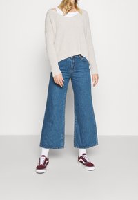 Beige stickad tröja med V-ringning och nedhasade axlar, tillsammans med vida blå denimjeans. Mörkröda sneakers med vita sulor fulländar outfiten.