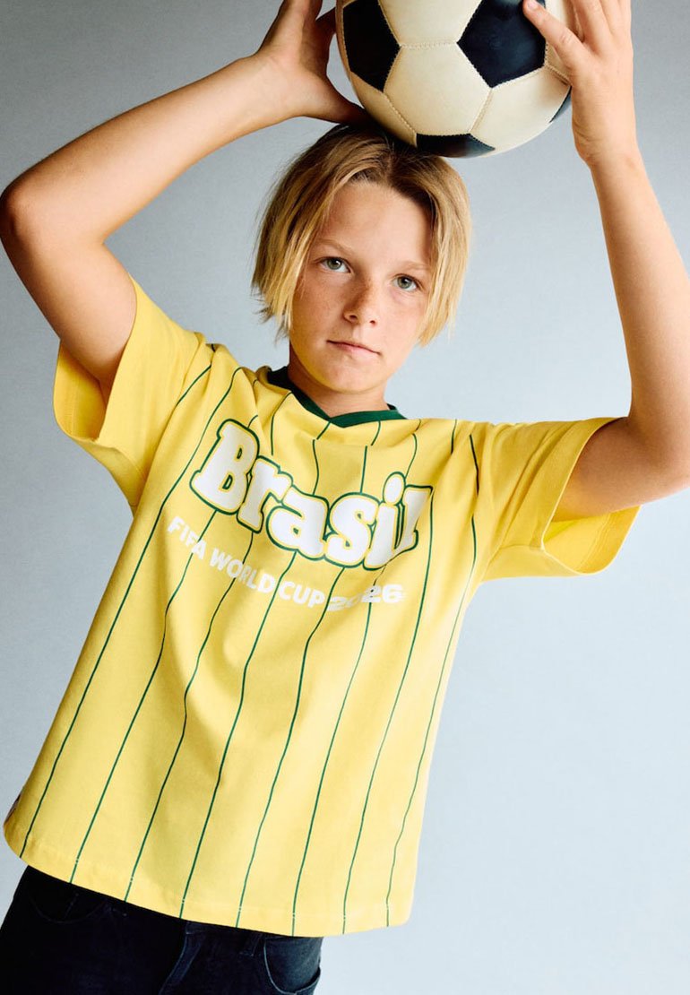 Jeune portant le maillot jaune du Brésil Coupe du Monde FIFA 2026 tenant un ballon de football classique noir et blanc au-dessus de sa tête.