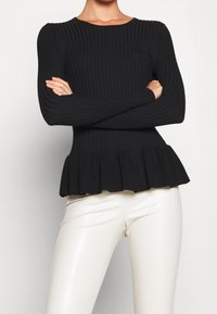 Black ribbed peplum top with long sleeves and a round neckline, featuring a flared hem. Paired with fitted white pants.