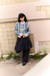 Cardigan tricotat cu modele geometrice albastre și verzi, guler crem; fustă cu carouri negre; pantofi negri uni; design minimal.