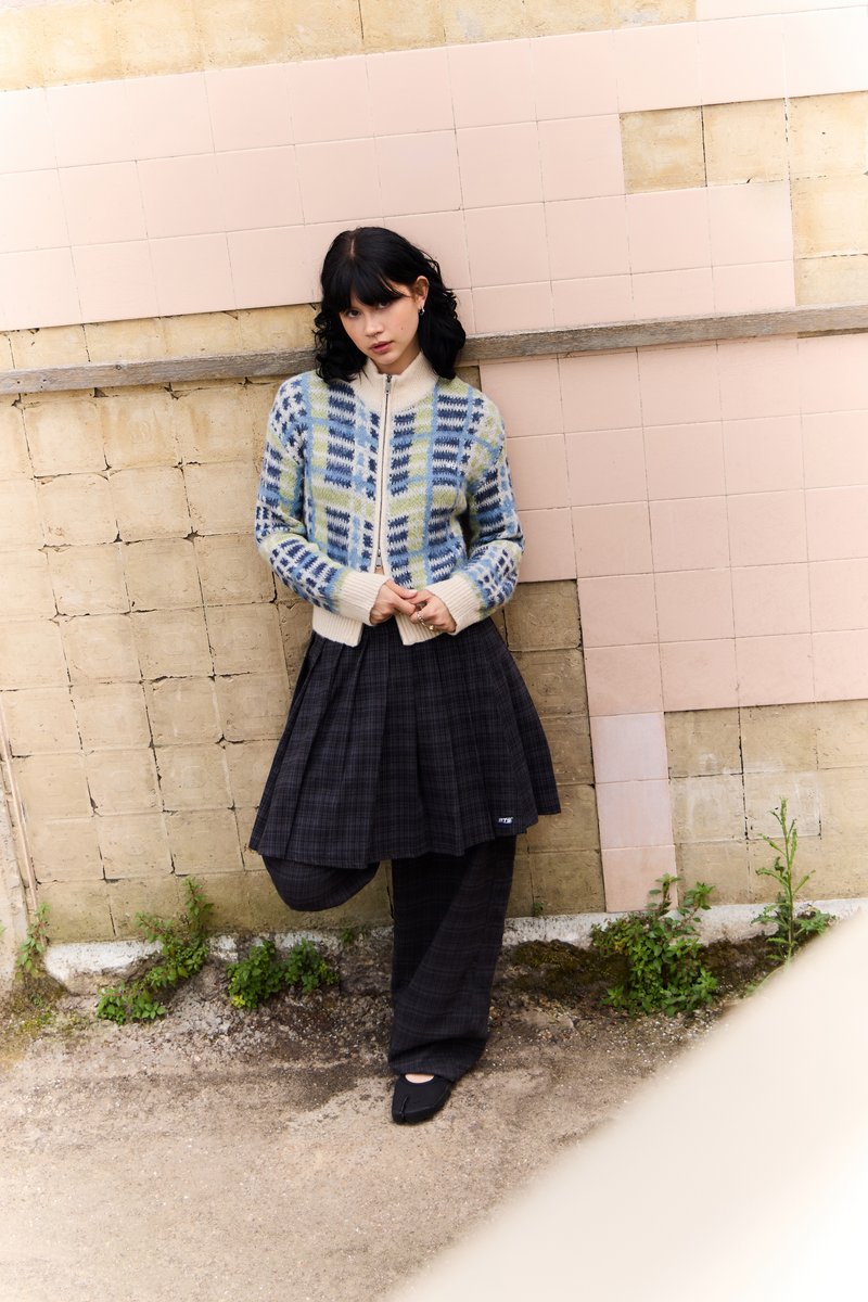 Cardigan tricotat cu modele geometrice albastre și verzi, guler crem; fustă cu carouri negre; pantofi negri uni; design minimal.