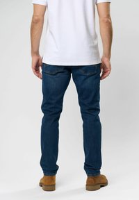 Sötétkék farmernadrág, slim fit szabással, hátsó zsebekkel és narancssárga varrással. Fehér inggel és barna cipővel viselve.