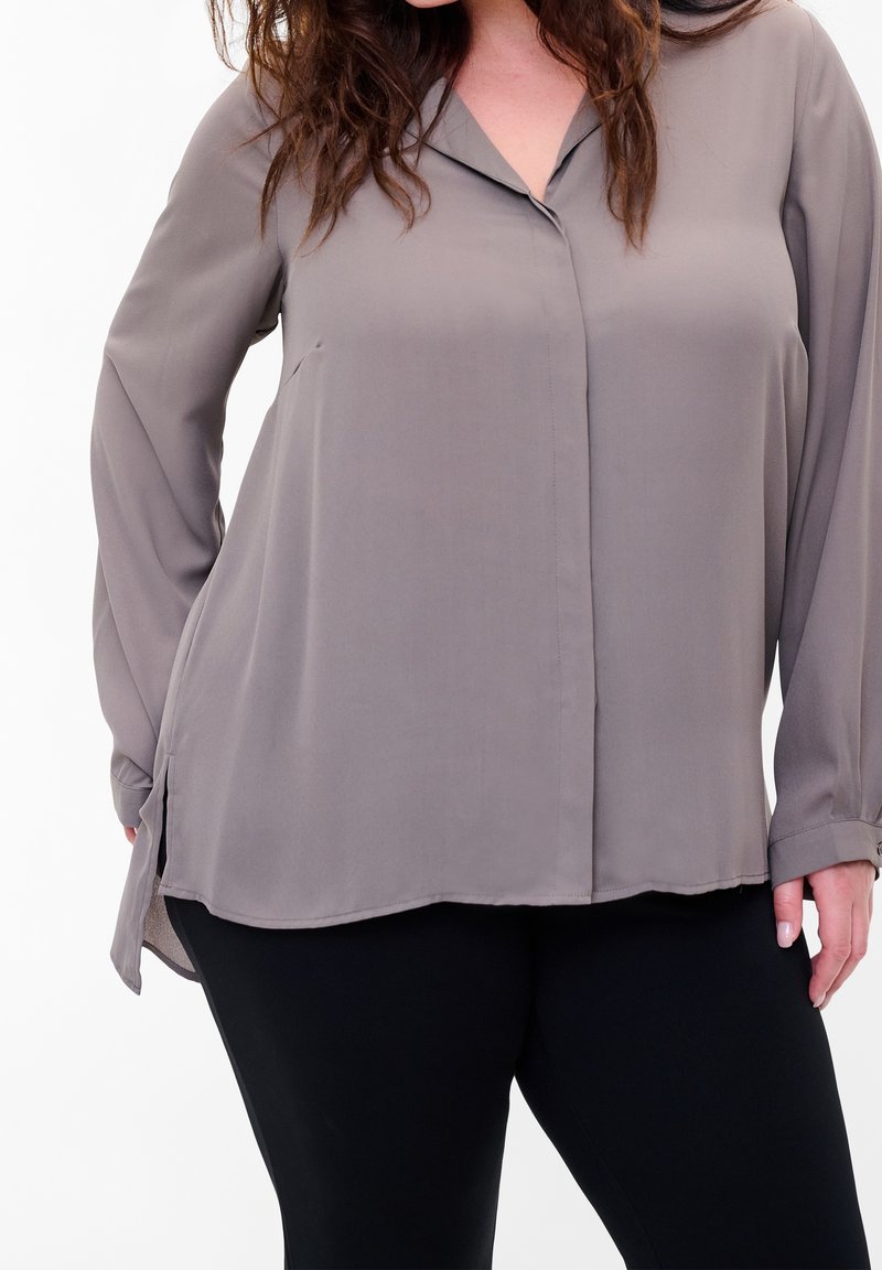 Femme portant un chemisier taupe ample à manches longues et un pantalon noir, debout devant un fond blanc uni.