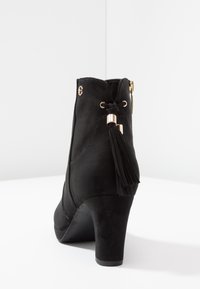 Černá semišová kotníková bota s vysokým podpatkem, která má šněrování, zlaté kovové prvky a dekorativní třapec na zadní straně.