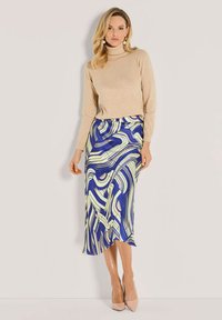 Beiger Rollkragenpullover kombiniert mit einem langen blauen Rock, der abstrakte grüne und weiße Muster aufweist. Model in hautfarbenen High Heels.