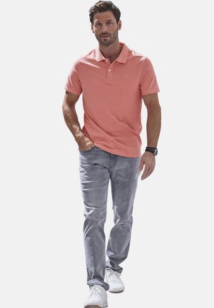 Mann geht selbstbewusst und trägt ein korallenrotes Polohemd, hellgraue Jeans, weiße Sneakers und eine Armbanduhr am linken Handgelenk.