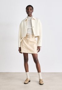 Veste blanche courte avec une texture côtelée, jupe en cuir beige à taille ceinturée, top crème texturé, et mocassins couleur beige avec des chaussettes.