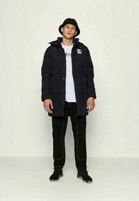 Marinblå pufferjacka med huva och logotyp, knappslutning fram, två sidofickor, bärs över en vit T-shirt och svarta byxor med vita detaljer.