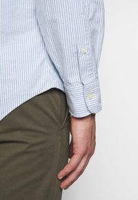 Blå och vitrandig skjorta med knappar och manschettdetalj, kombinerad med olivgröna byxor. Mjukt tyg med en avslappnad passform.