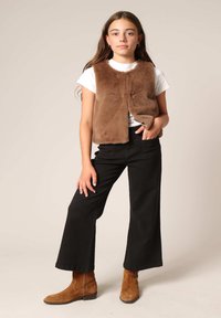 Gilet en fausse fourrure marron avec un col rond, associé à un t-shirt blanc et un pantalon noir à jambes larges. Des bottines en daim marron à la cheville complètent le look.