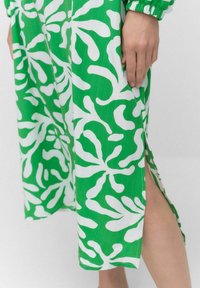 Vihreä mekko, jossa on valkoinen abstrakti lehtikuosi ja sivuaukko. Kankaalla on teksturoitu pinta ja elastiset hihansuut.