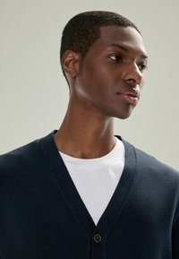 Ung mand iført en marineblå, knappet cardigan over en hvid crew neck skjorte, ser tankefuldt til siden mod en ensfarvet baggrund.