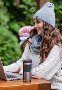 Grijs gebreid beanie en sjaal, gezellige romige kabelgebreide trui, metalen reisbeker en laptop op een houten tafel, met groenblijvende planten op de achtergrond.